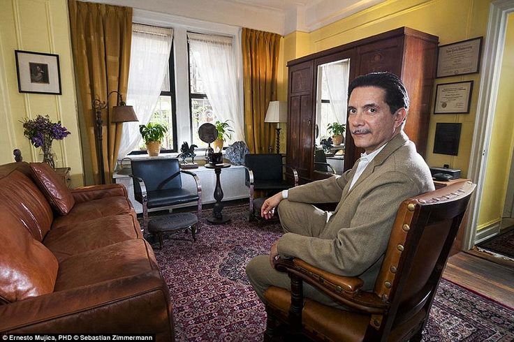 a man sitting in a living room next to a brown leather couch and chair on top of a rug