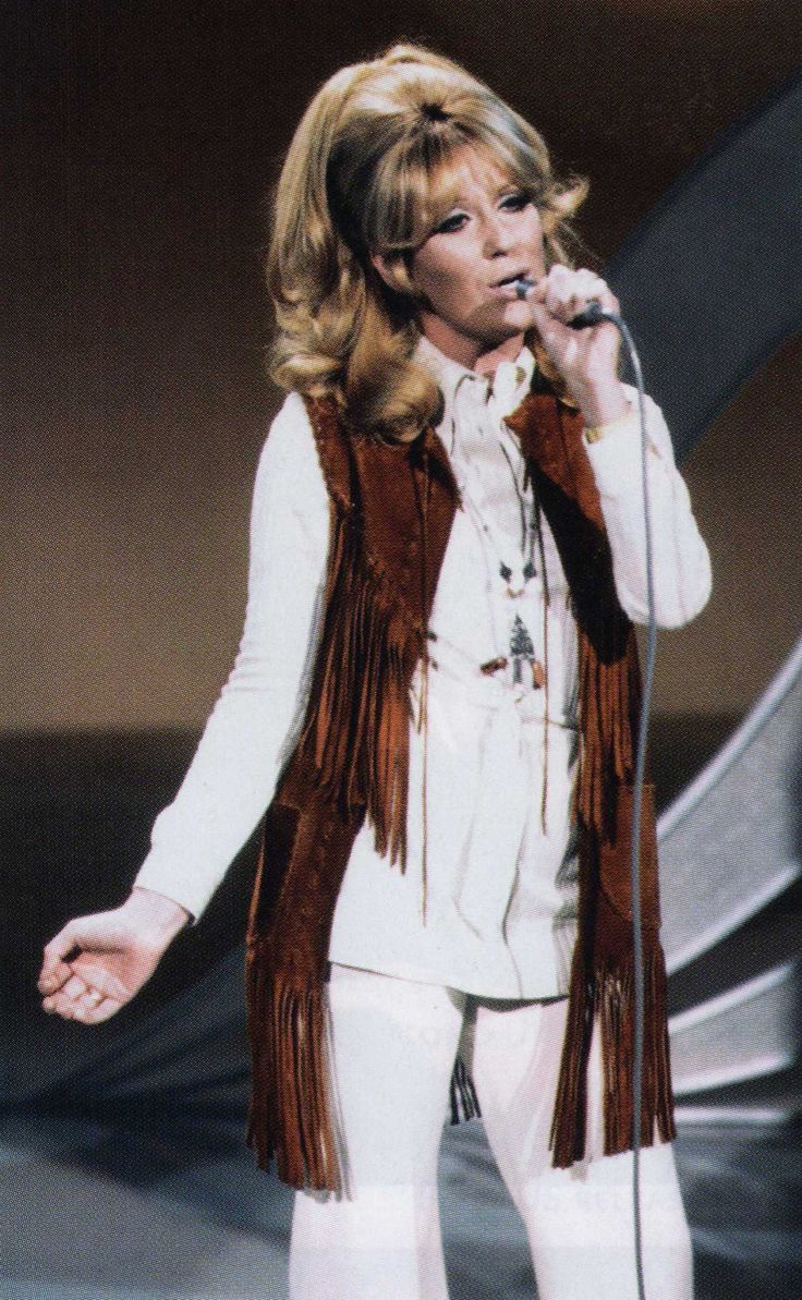 a woman in white pants and brown fringe vest singing into a microphone while standing on stage