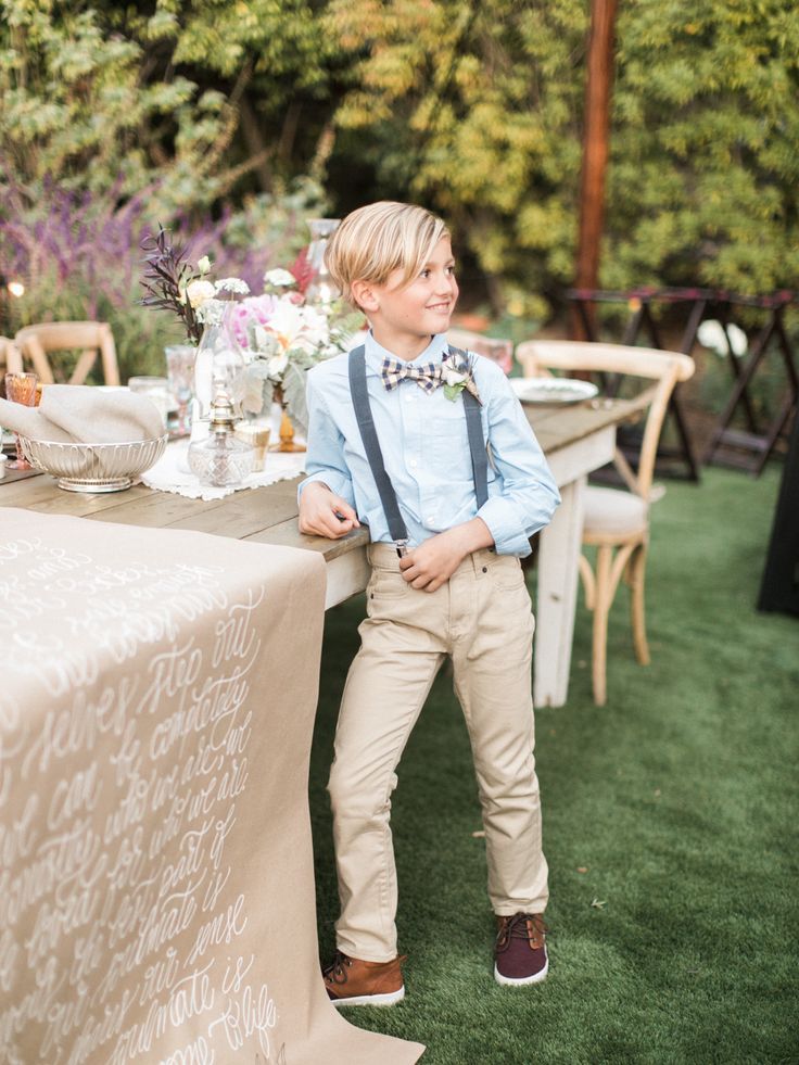 a young boy wearing suspenders and a bow tie standing in front of a table