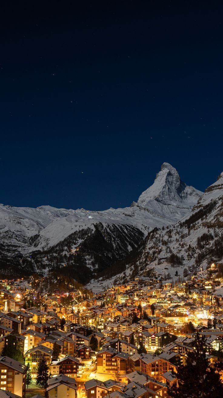 the city is lit up at night with mountains in the background and snow on the ground