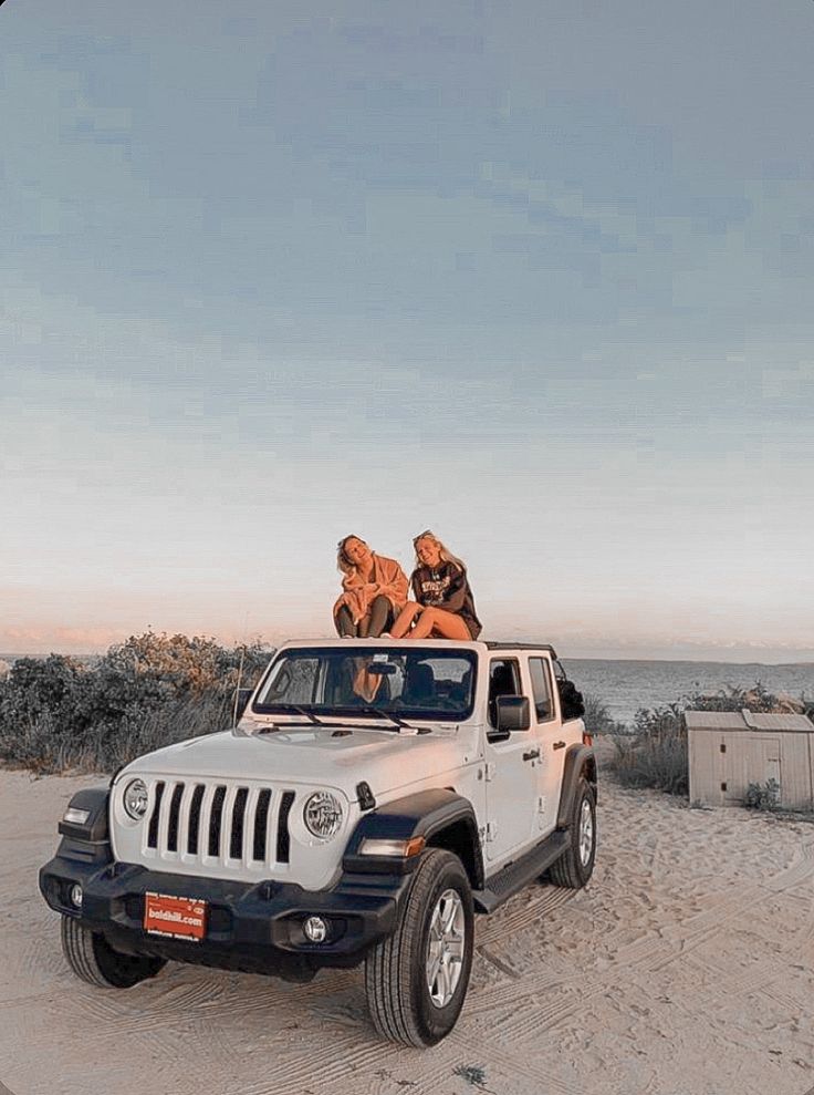 two people are sitting on top of a jeep in the sand at the beach,