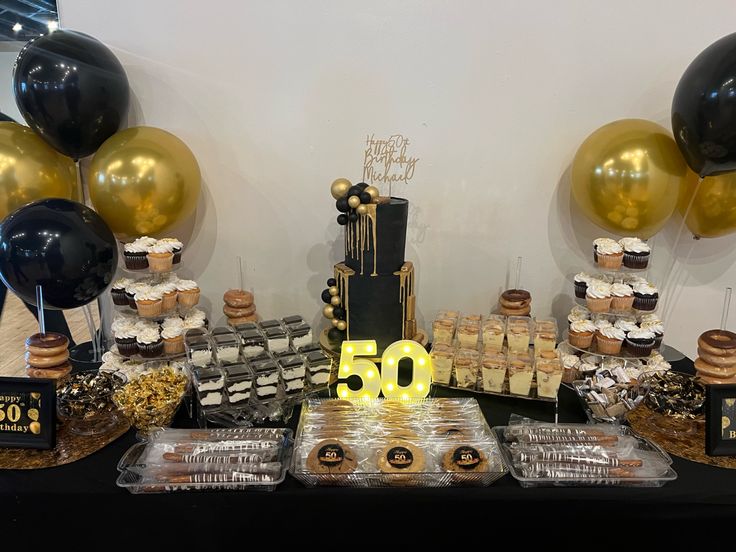 a table topped with lots of desserts next to black and gold balloons in the background