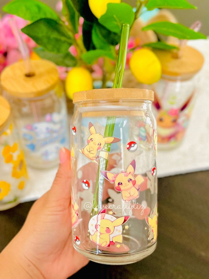 a hand holding a glass with pokemon characters on it and some flowers in the background