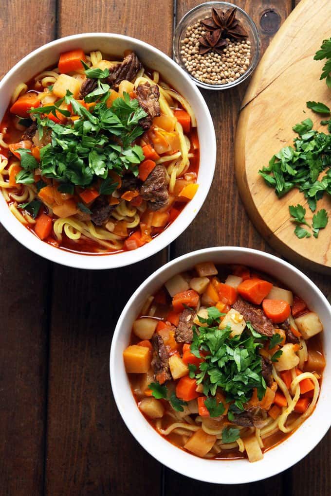 two bowls of soup with meat, carrots and parsley next to a cutting board