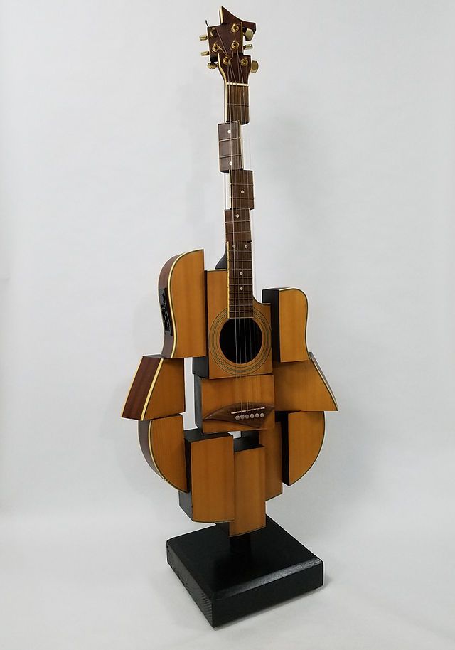 a wooden guitar sculpture sitting on top of a black stand next to a white wall