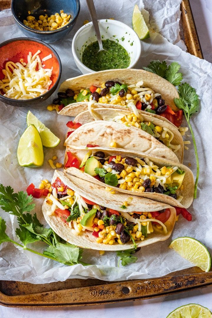 three tacos on a tray with salsa, corn and cilantro in the background