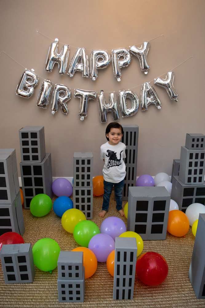 a young boy standing in front of a birthday cake surrounded by balloons and city buildings