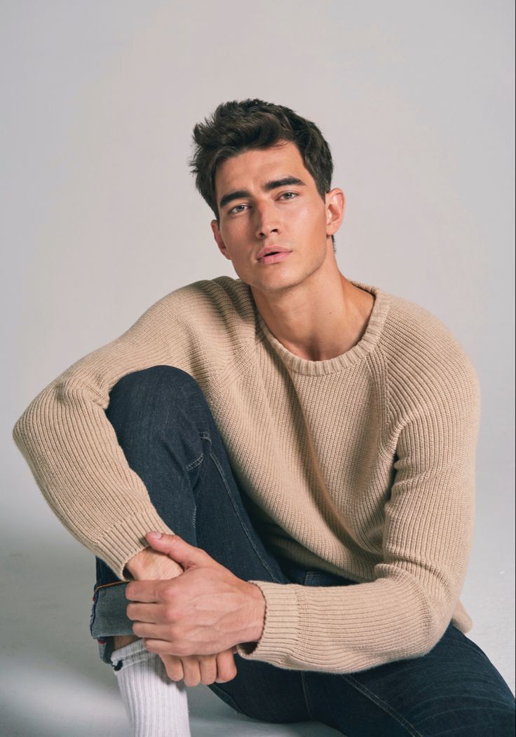 a young man sitting on the ground wearing a sweater and jeans with his legs crossed