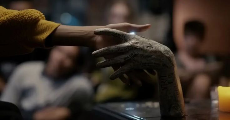 two hands reaching for a candle on a table with other people in the room behind them