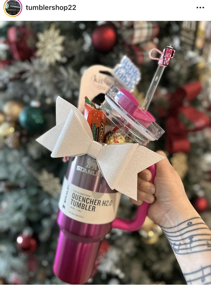 someone is holding up a pink cup with a bow on it in front of a christmas tree