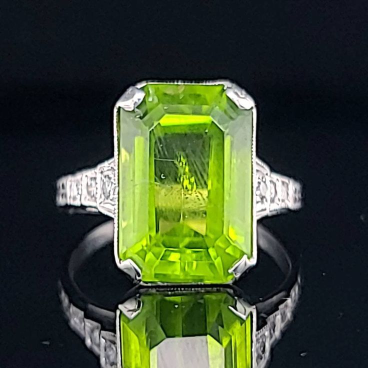 an emerald colored ring with diamond accents on it's sides, sitting on a black surface