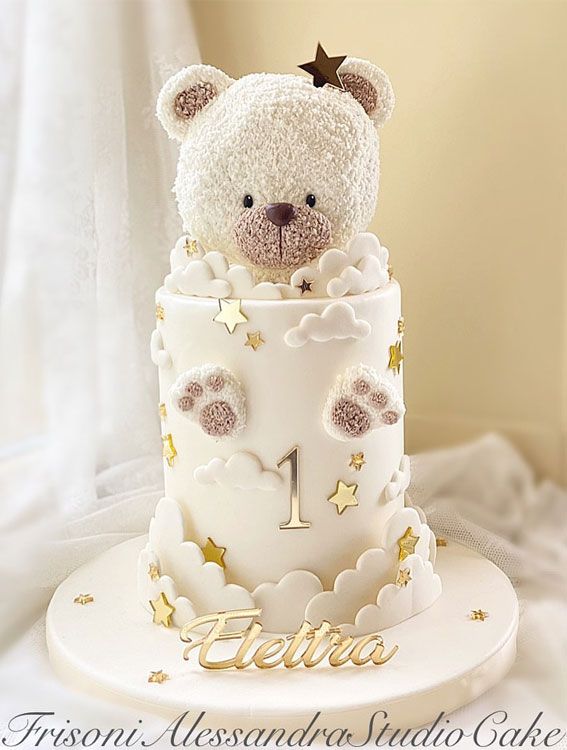 a teddy bear sitting on top of a white cake with gold stars and clouds around it