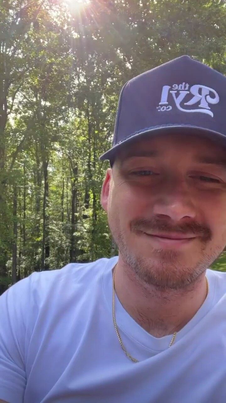 a man wearing a blue hat and white shirt in front of some trees with the sun shining on him