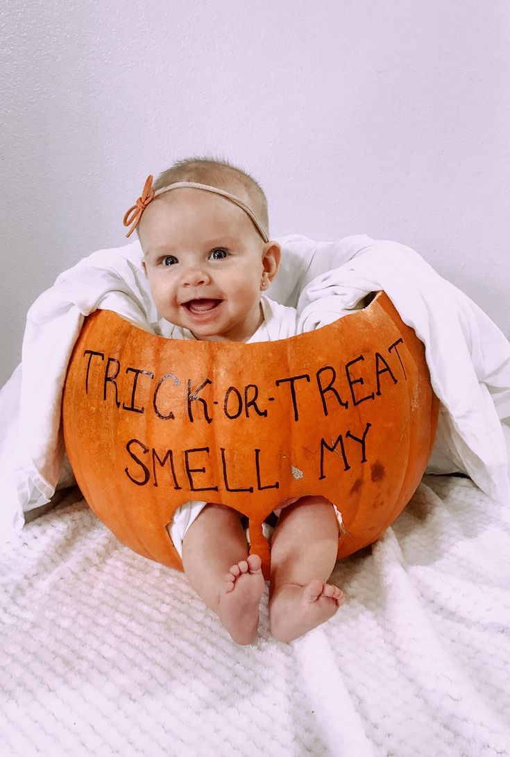 a baby sitting on top of a bed with a pumpkin shaped pillow that says trick or treat smell my