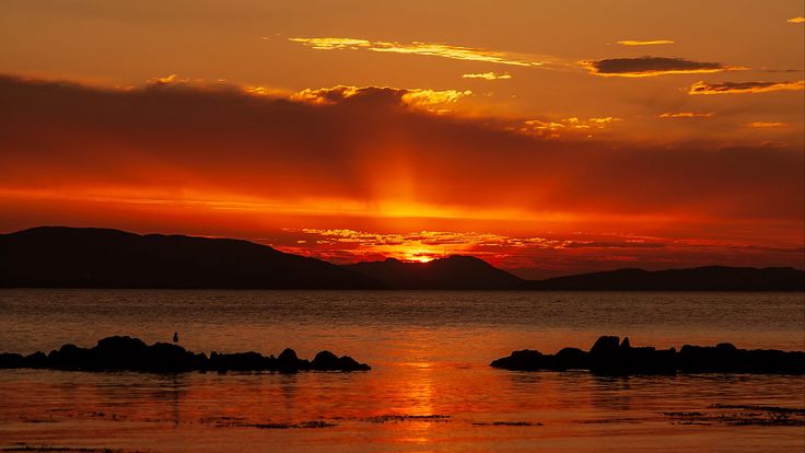 the sun is setting over the ocean with rocks in the foreground and mountains in the distance