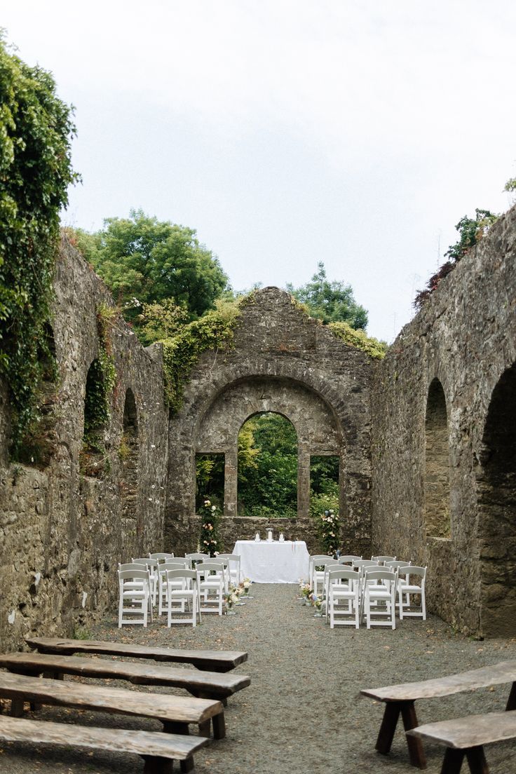 an outdoor ceremony setup with white chairs and flowers on the aisle, surrounded by stone archways