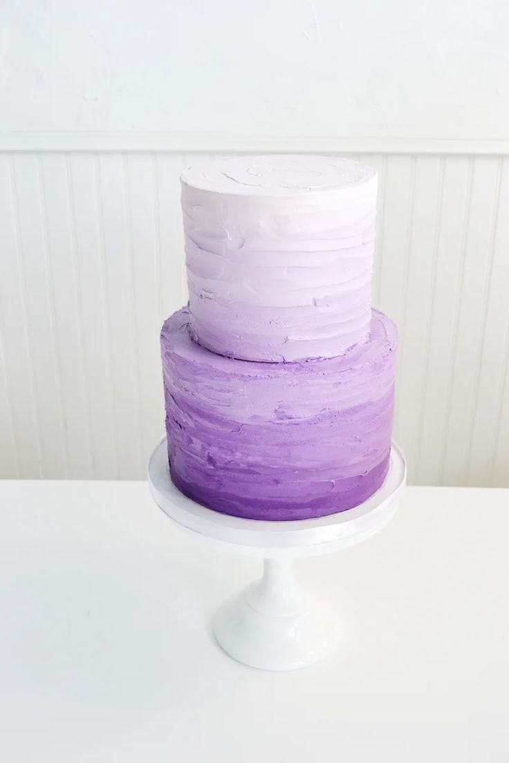 a three layer cake with purple icing on a white pedestal in front of a striped wall