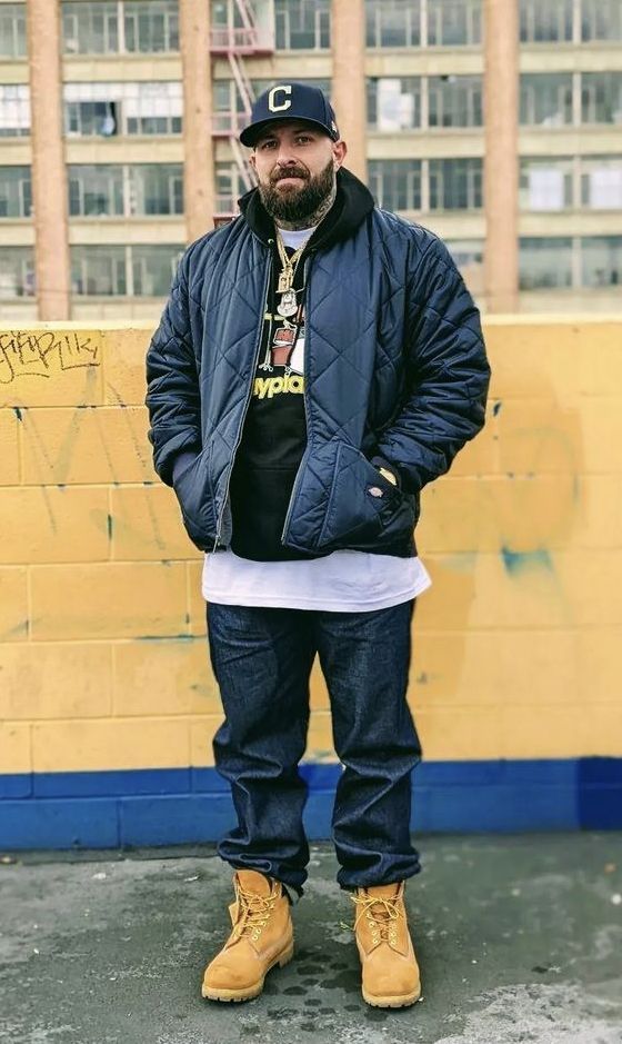 a man standing in front of a yellow wall wearing a black jacket and blue jeans