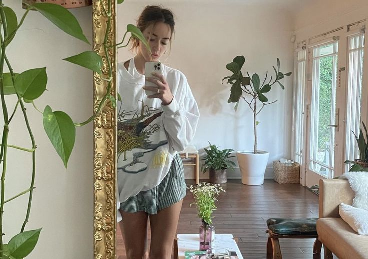 a woman standing in front of a mirror looking at her cell phone while holding a plant