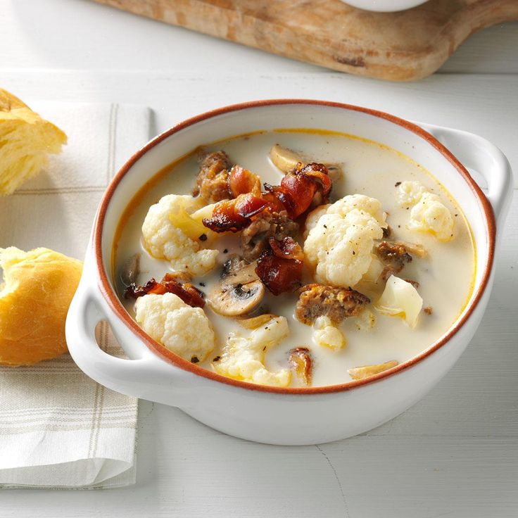 a bowl of soup with cauliflower and bacon in it next to bread on a cutting board