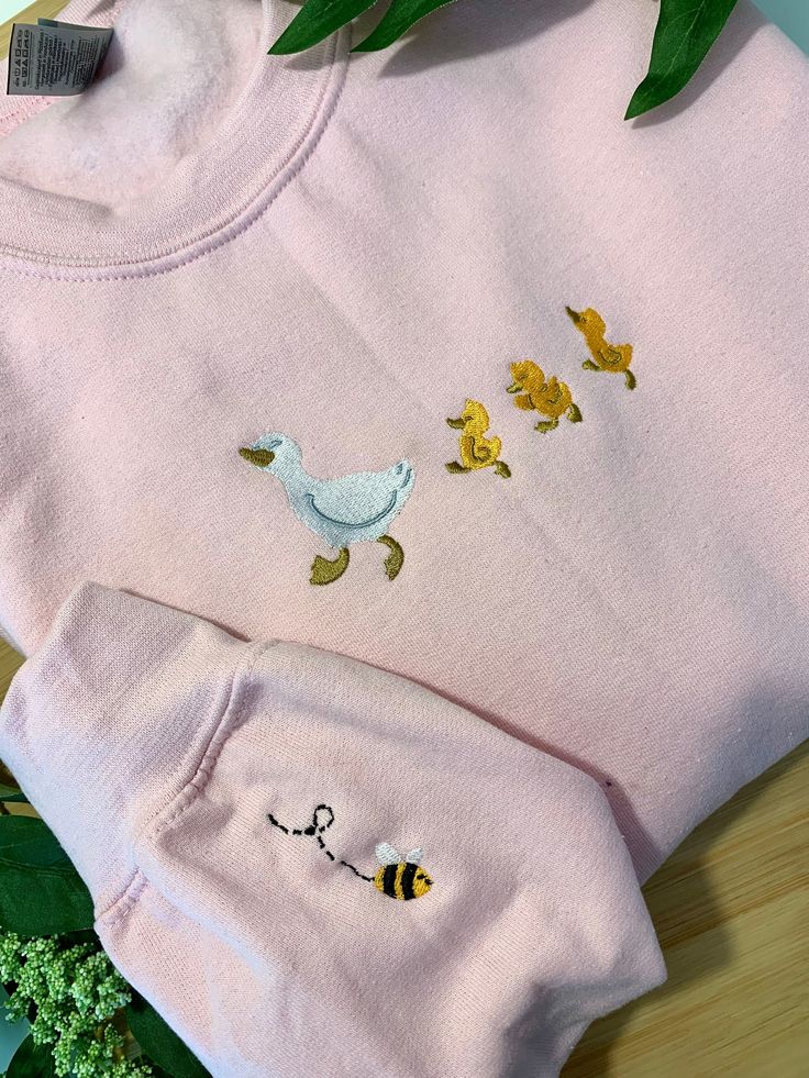 two pink sweaters with embroidered ducks and bees on them, sitting next to broccoli