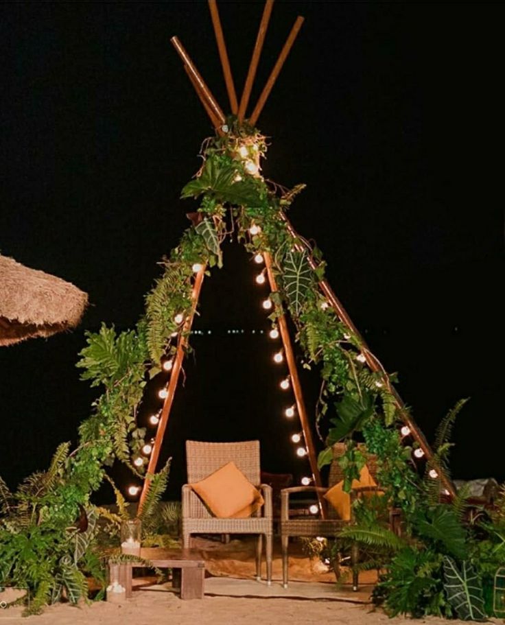 a teepee tent decorated with lights and greenery for an outdoor seating area at night