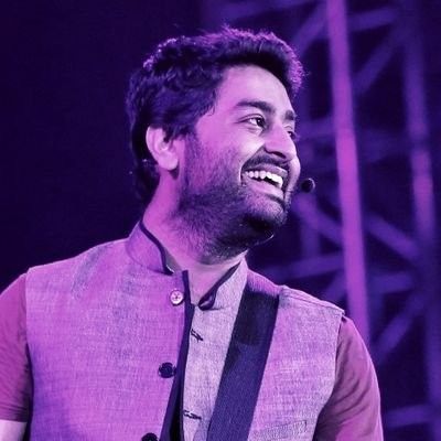 a man with a guitar in his hand and wearing a purple vest, smiling at the camera