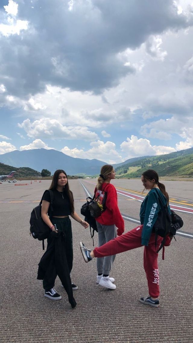 three girls are standing in the middle of an airport tarmac with mountains in the background