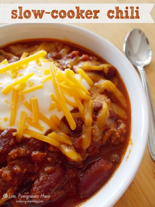 a bowl filled with chili and cheese on top of a wooden table next to a spoon