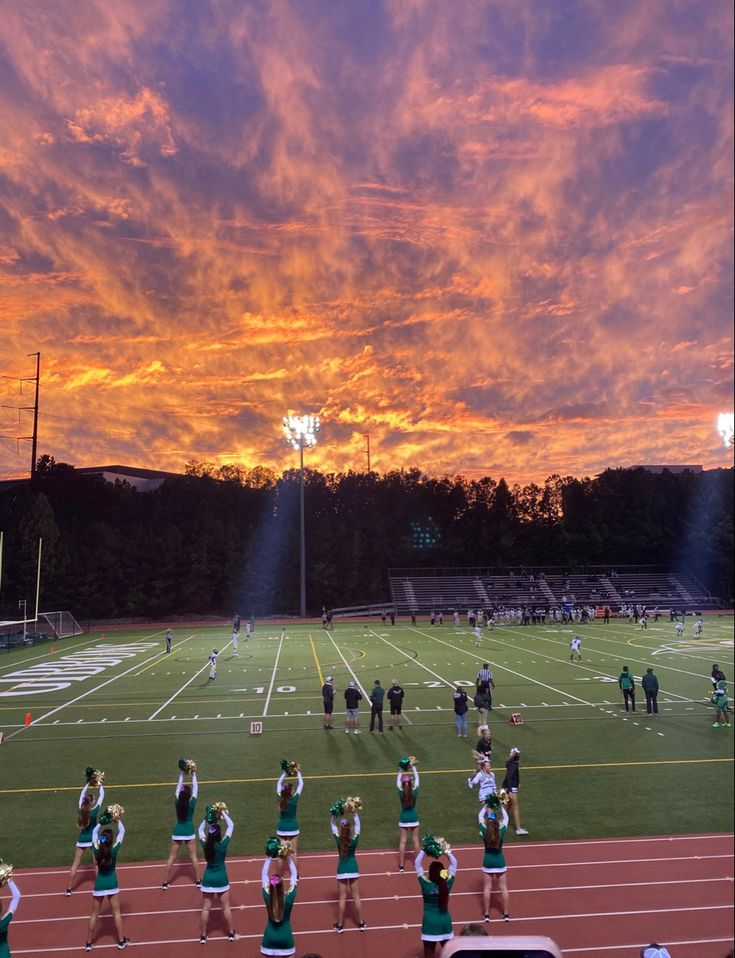 a group of cheerleaders on the sidelines at a football game under a colorful sky