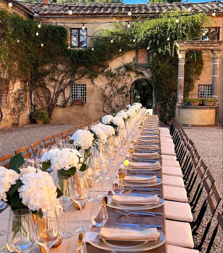 a long table is set with white flowers and place settings for an outdoor dinner party