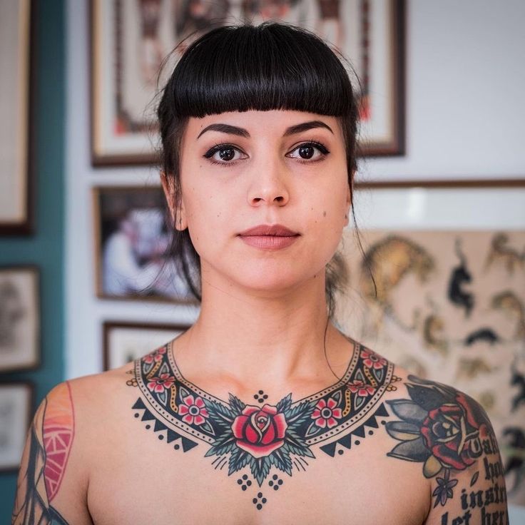 a woman with tattoos on her chest looking at the camera while standing in front of pictures