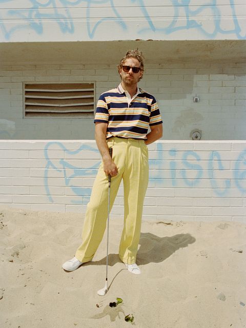 a man in striped shirt and yellow pants holding a golf club on sand with graffiti behind him