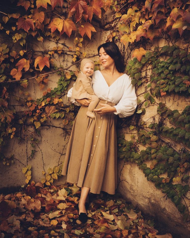 a woman holding a baby standing in front of a wall covered with vines and leaves