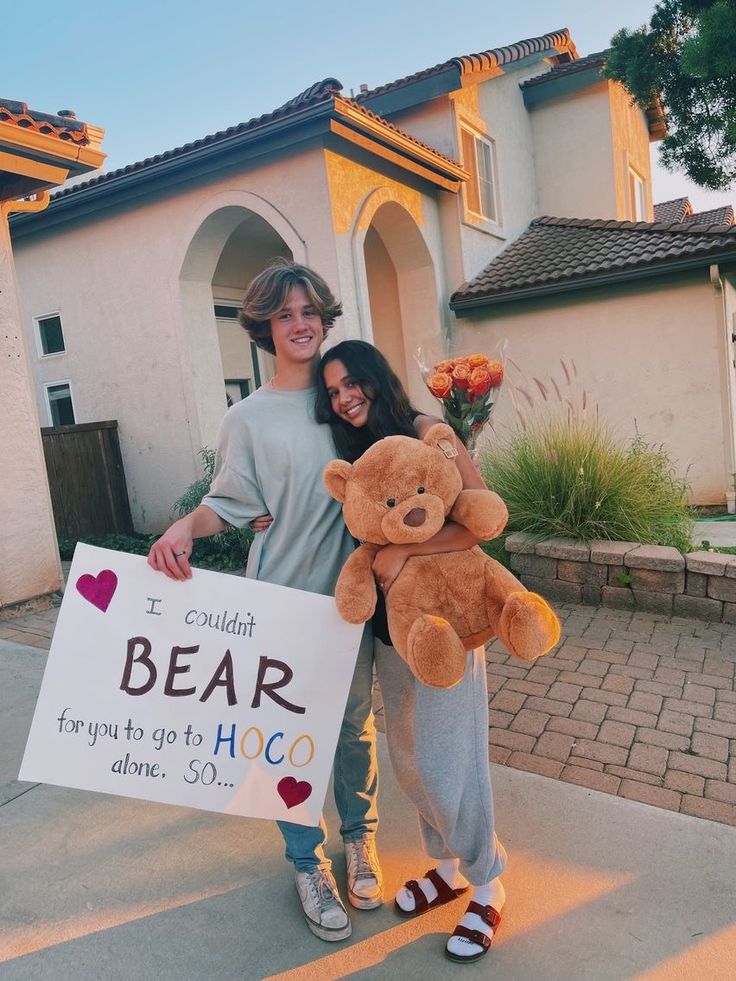 two people standing in front of a house holding a teddy bear and a sign that says i couldn't bear you got to hoo