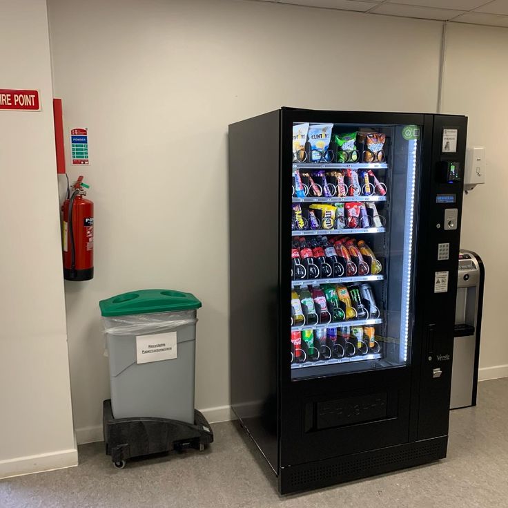 a vending machine next to a trash can in an empty room with white walls