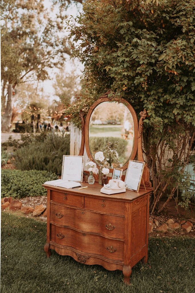 an old dresser with a mirror and flowers on it in front of a large tree