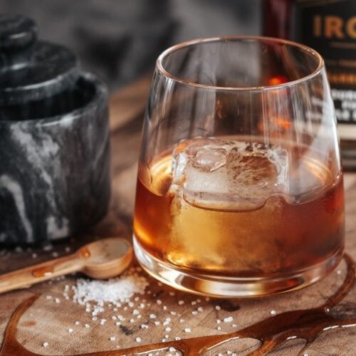 an old fashioned glass filled with ice sits on a wooden table next to a bottle and spoon