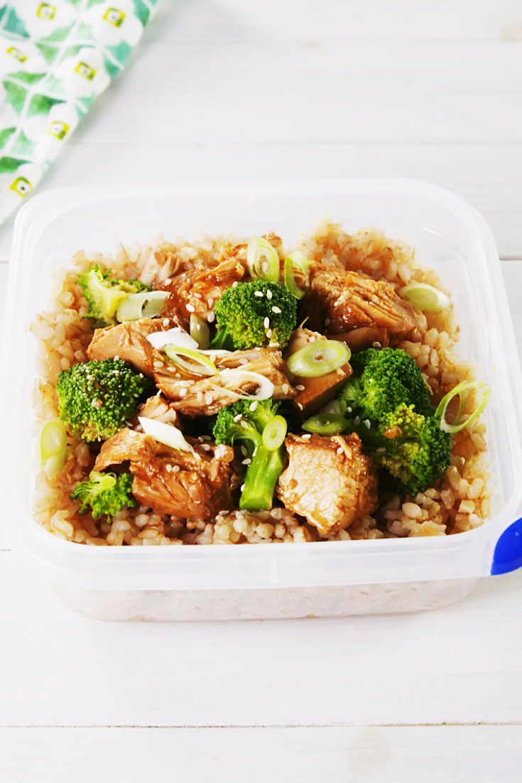 a plastic container filled with chicken and broccoli on top of brown rice next to a blue spoon