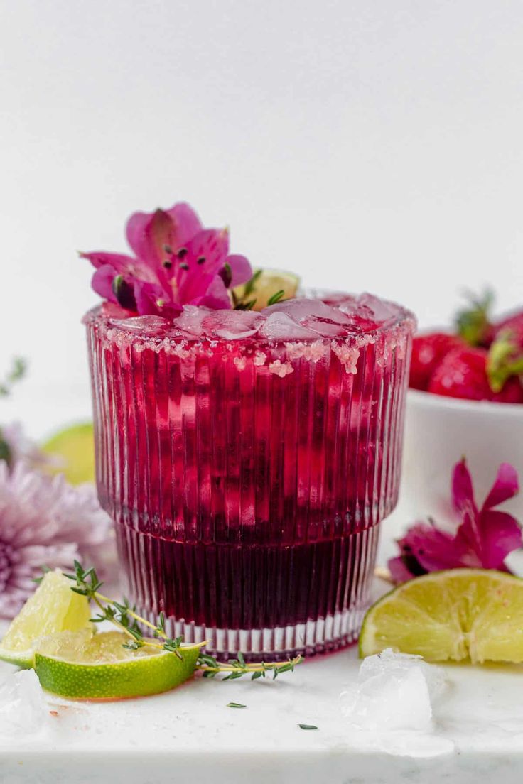 a red drink in a glass with flowers and limes around it on a white surface