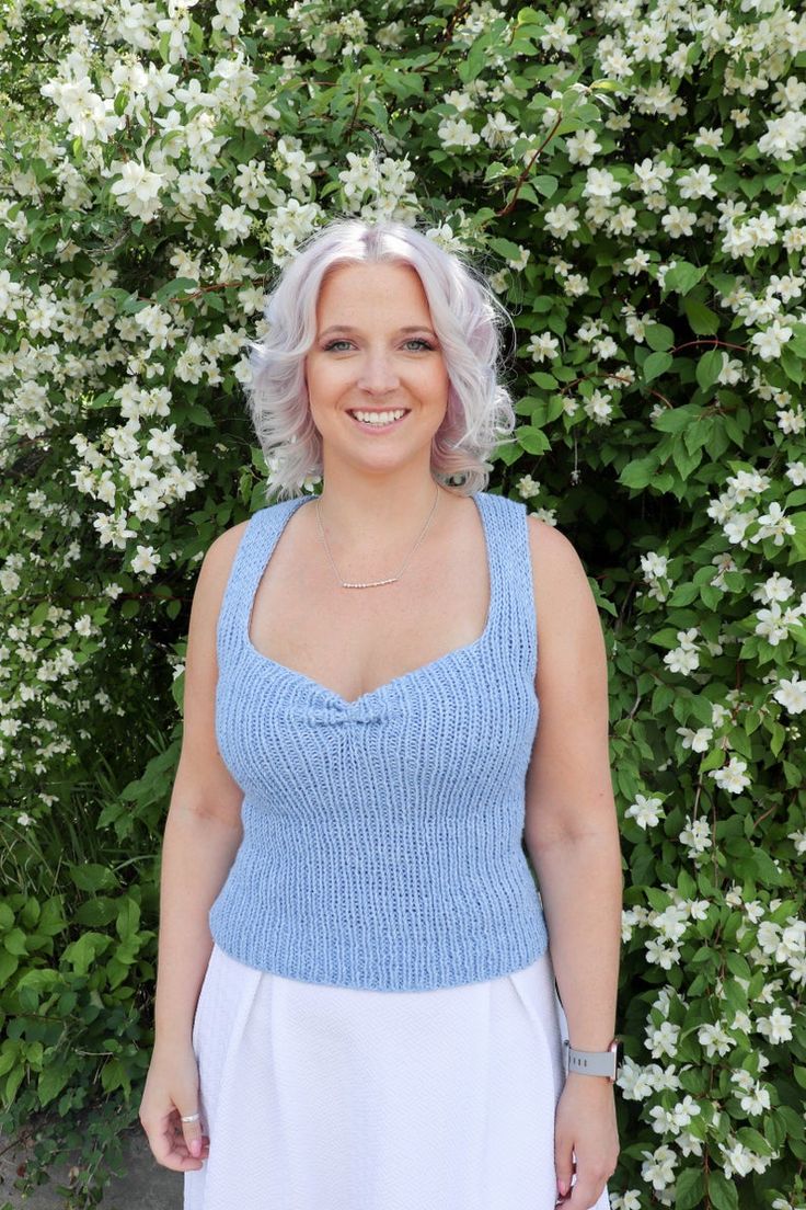 a woman with white hair wearing a blue knitted top standing in front of flowers