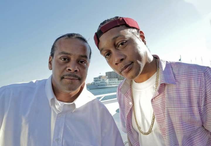 two men standing next to each other in front of a body of water with boats in the background