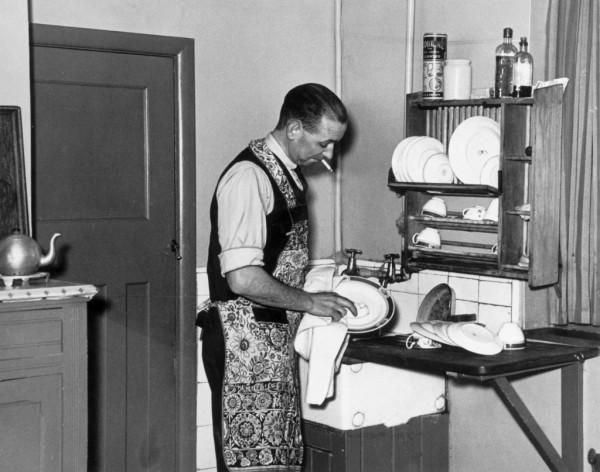 an old black and white photo of a man ironing clothes in his kitchen,