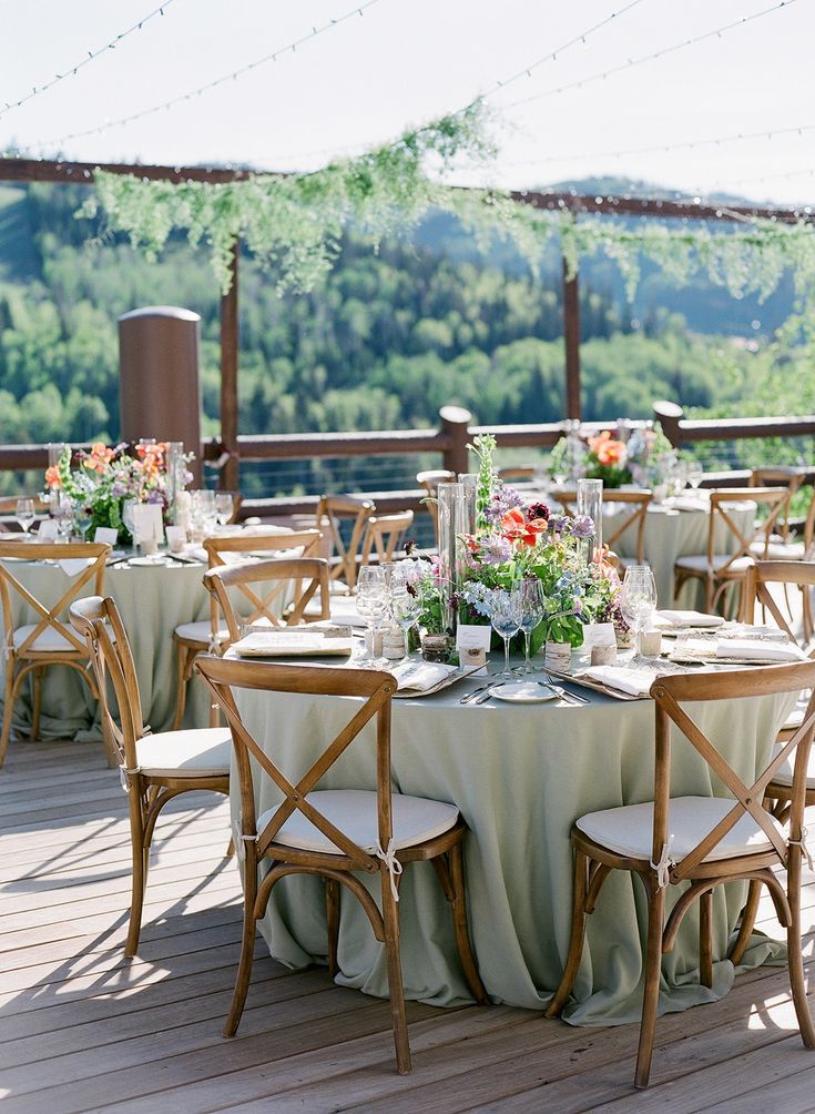 an outdoor table set up with flowers and greenery on the top of a wooden deck