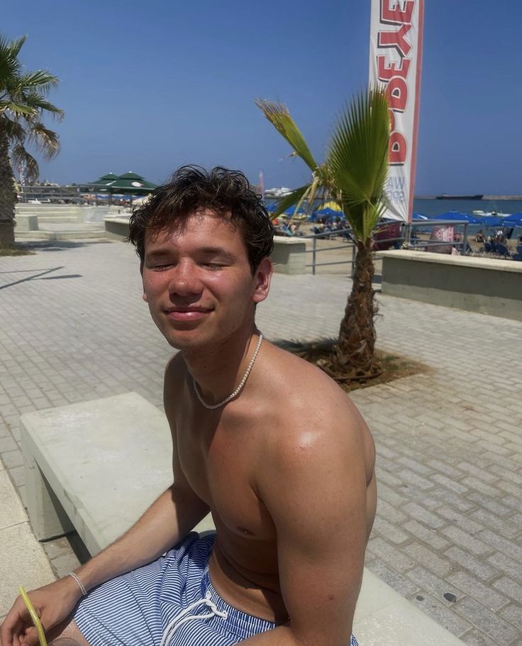 a shirtless man sitting on a bench next to the ocean with his eyes closed