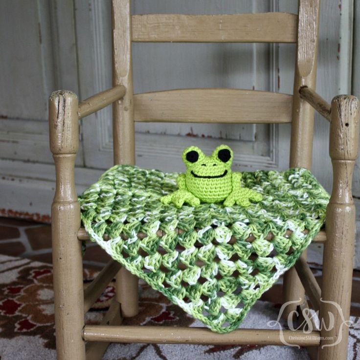 a crocheted frog sitting on top of a green blanket in a wooden rocking chair