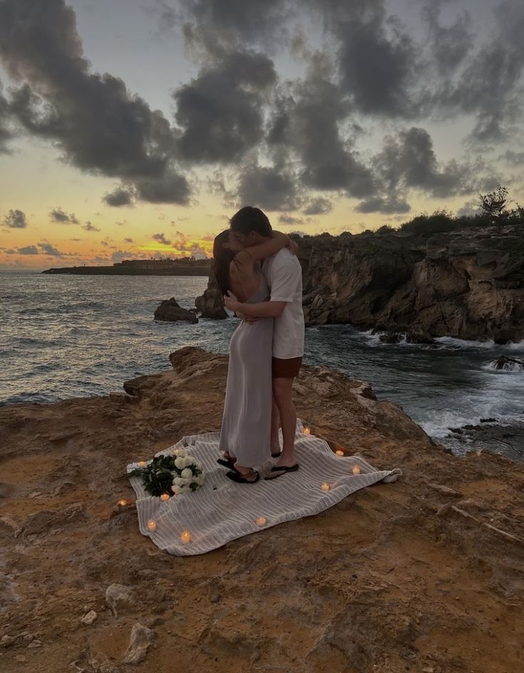 a man and woman standing on top of a rock next to the ocean at sunset