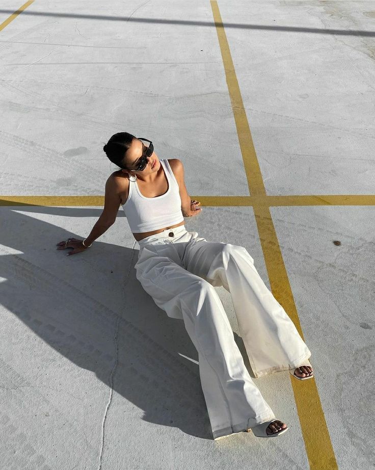 a woman sitting on the ground wearing white pants and a tank top with her hands in her pockets