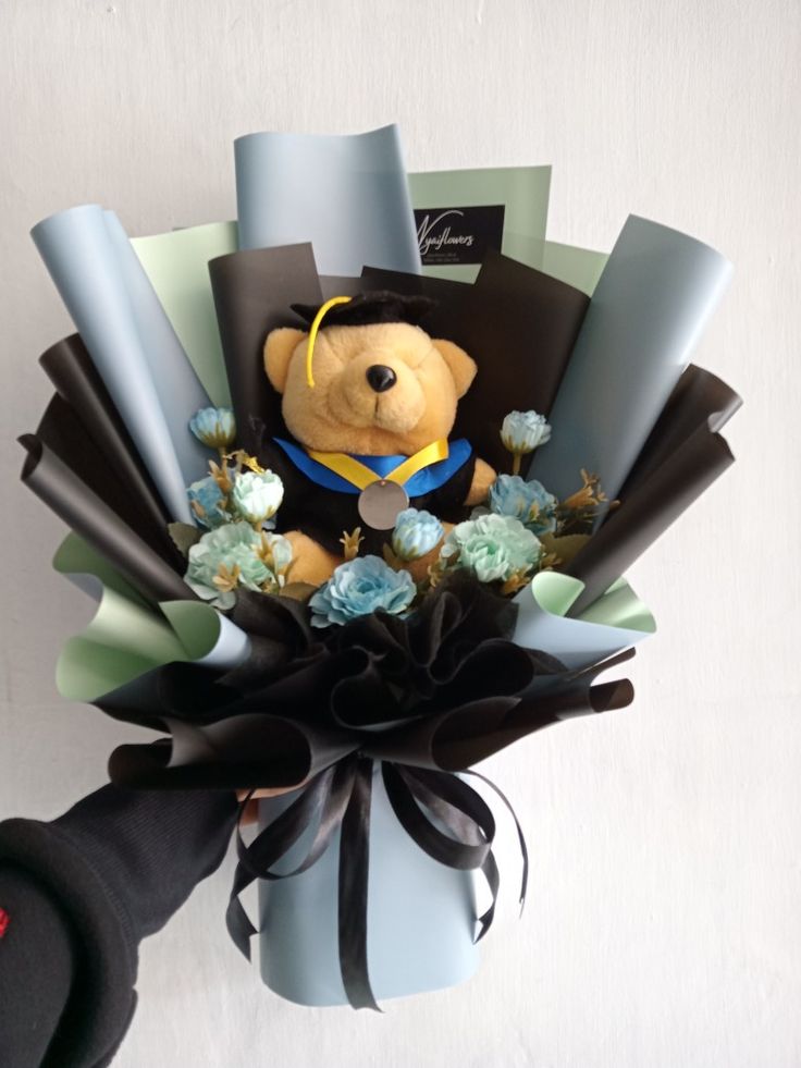 a teddy bear in a graduation cap and gown surrounded by blue flowers, black ribbon, and other items