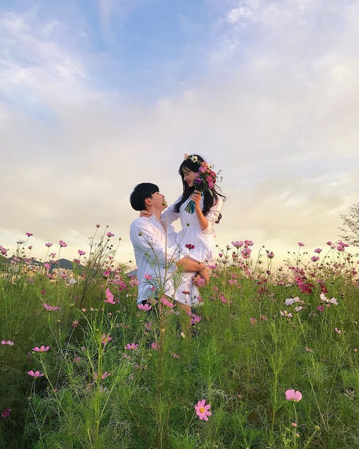 a man and woman standing in the middle of a field with pink flowers on it